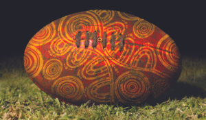 An AFL Football on grass with illustrations on the ball by Murrupuk Art